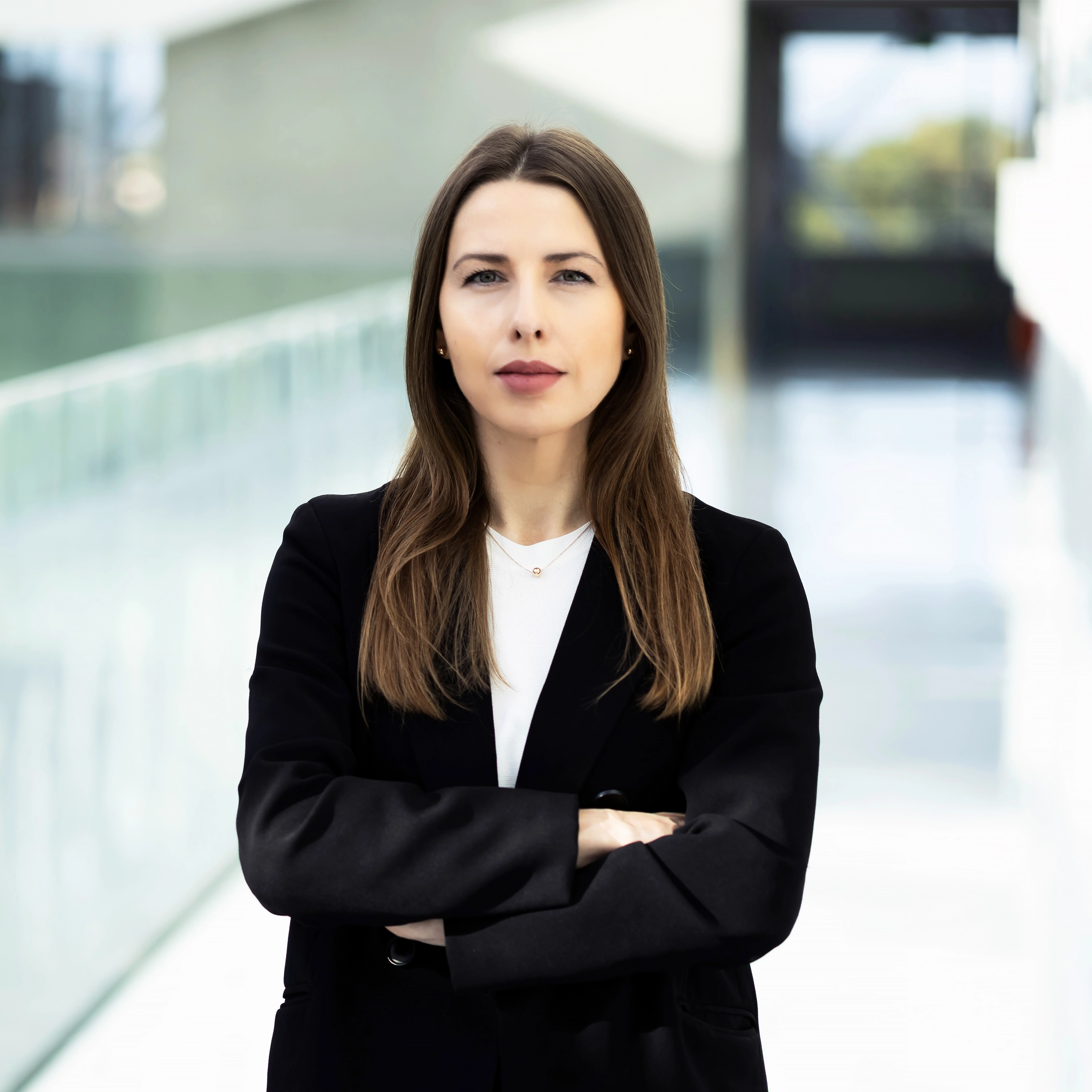 A young woman with straight light brown hair and a black blazer stands in a modern glass corridor, arms crossed, looking confidently into the camera.