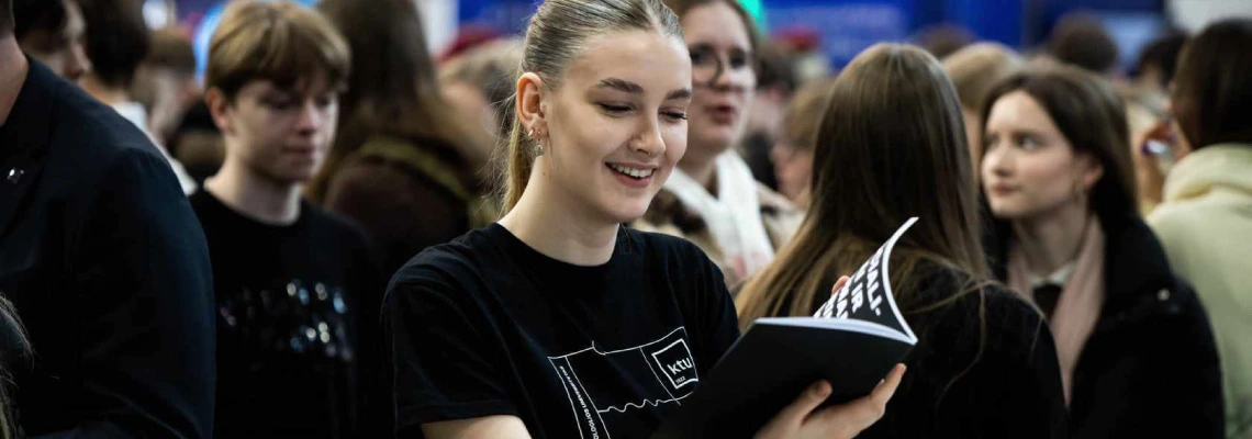 A young woman with tied-back hair and a black T-shirt featuring the "KTU" logo smiles while looking at an open booklet or magazine. She is surrounded by a crowd of mostly young people, suggesting the setting is a public event or fair.