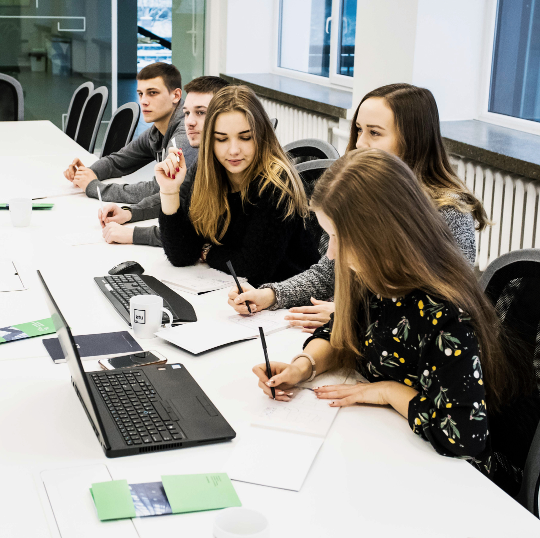 KTU students working together at a shared table, discussing tasks, writing notes and using laptops in a university learning environment.