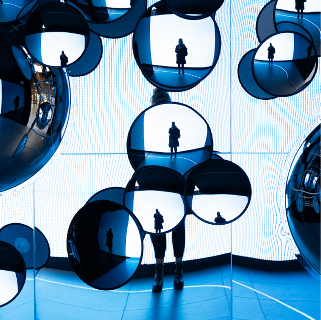 A person standing among multiple reflective circular mirrors in an immersive blue-lit installation, with their silhouette repeated across the reflections, representing the strategic direction &ldquo;Engaging and Creative Community&rdquo;.