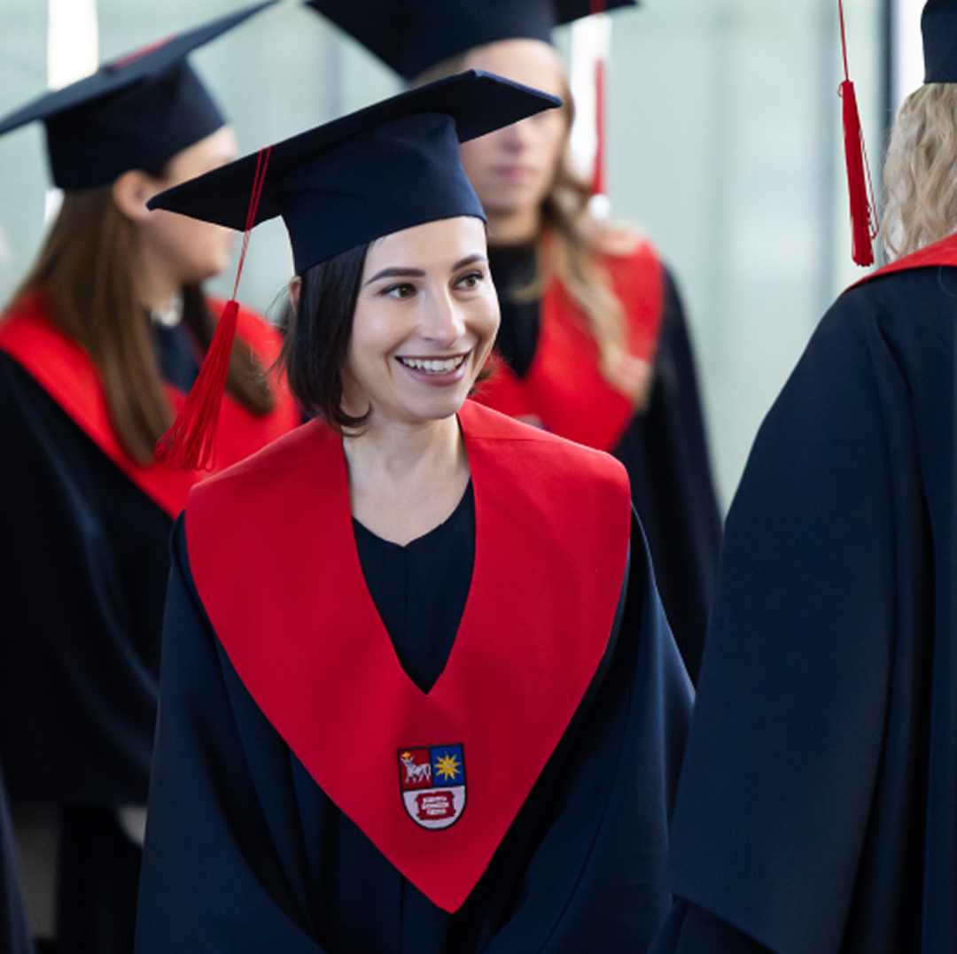 A smiling KTU graduate wearing academic robes, a graduation cap and a red stole, surrounded by other graduates during a graduation ceremony, representing the strategic direction &ldquo;Engaging Transformative and Empowering Learning for the Future&rdquo;.