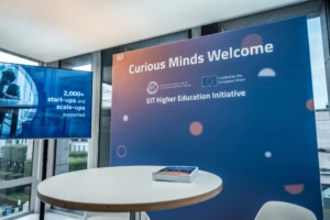 A conference welcome area with a large blue EIT Higher Education Initiative banner reading “Curious Minds Welcome,” alongside EIT and European Union funding logos. A nearby digital screen says “2,000+ start-ups and scale-ups supported,” with a round table and brochures in the foreground.