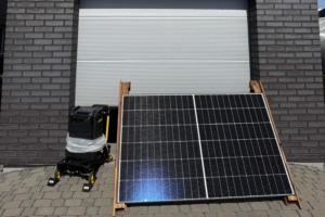 A portable solar power setup displayed outdoors in front of a gray brick building. A large solar panel is mounted on a wooden stand, with a portable battery power station on a small wheeled cart beside it. A closed metal roller door forms the background, and the equipment sits on a paved surface in bright sunlight.