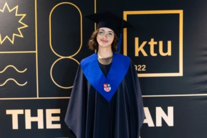 A smiling graduate wearing glasses, a dark academic gown, blue stole, and mortarboard poses in front of a black-and-gold KTU graduation backdrop.