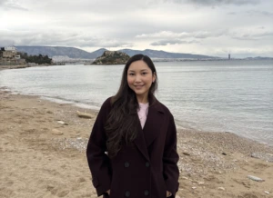 A woman with long dark hair stands smiling on a quiet sandy beach, wearing a dark burgundy coat over a light pink top. Calm water, a small rocky islet, distant mountains, and a cloudy sky create a peaceful coastal backdrop.