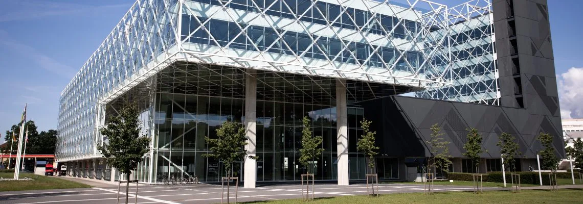 A modern glass building features a large transparent wall reflecting sunlight and the surrounding scenery represents nine KTU Faculties.