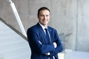 Man in a blue suit and tie standing with his arms folded, smiling at the camera in a bright, modern interior
